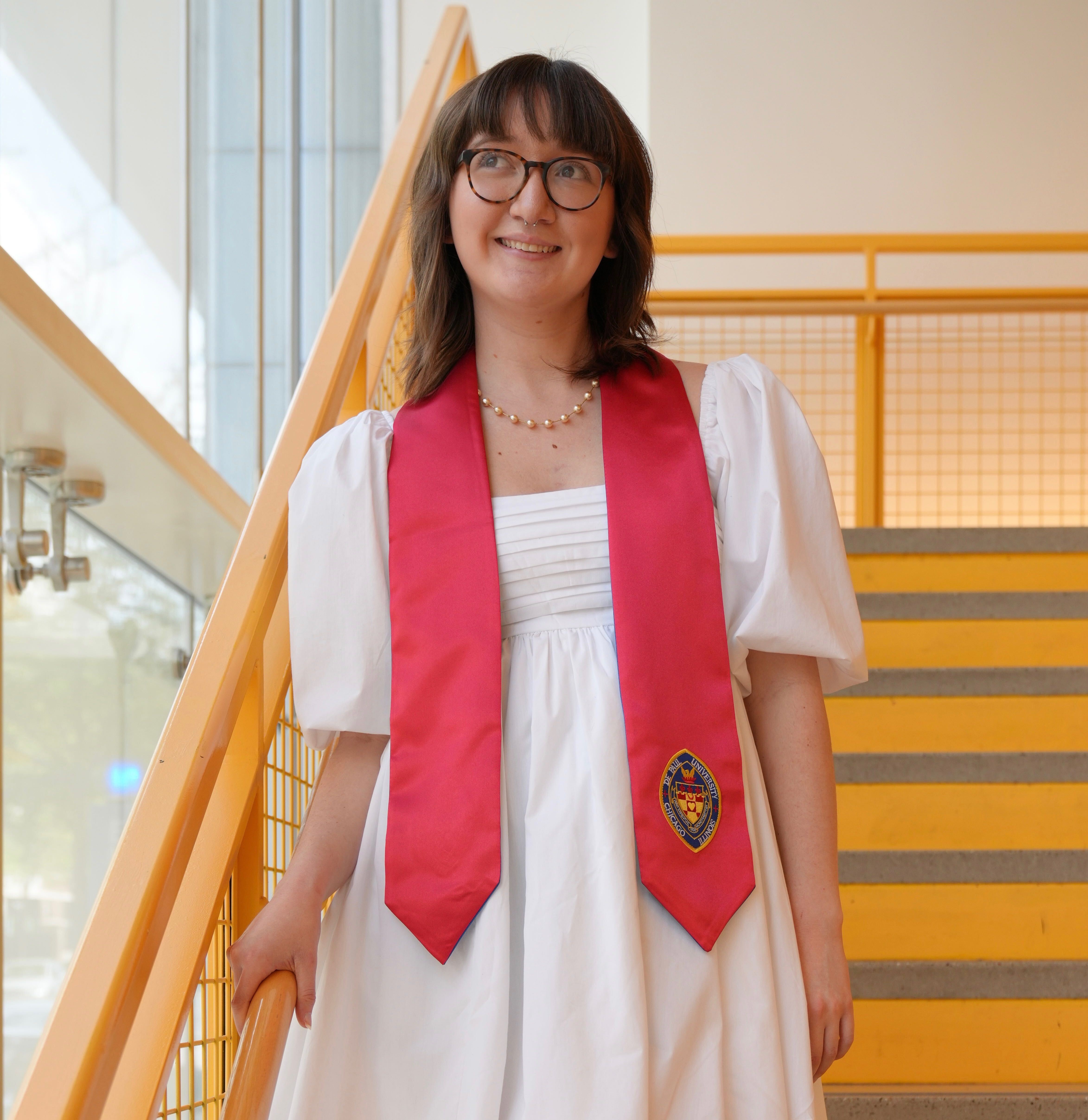Grace Archer stands on the yellow stairs and wears a white dress with a red graduation sash